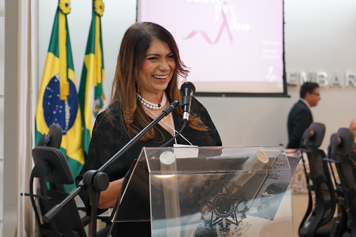 Uma mulher de meia-idade, com cabelos castanhos longos e um sorriso aberto, está em primeiro plano, atrás de um púlpito de acrílico transparente. Ela veste uma blusa preta e um colar de pérolas, e está com as mãos apoiadas no púlpito, onde há uma insígnia oficial e documentos. Um microfone está ajustado à sua frente. Ao fundo, à esquerda, duas bandeiras do Brasil estão lado a lado. Um homem, de terno escuro, está desfocado e de costas no lado direito, indicando um ambiente de evento ou conferência.