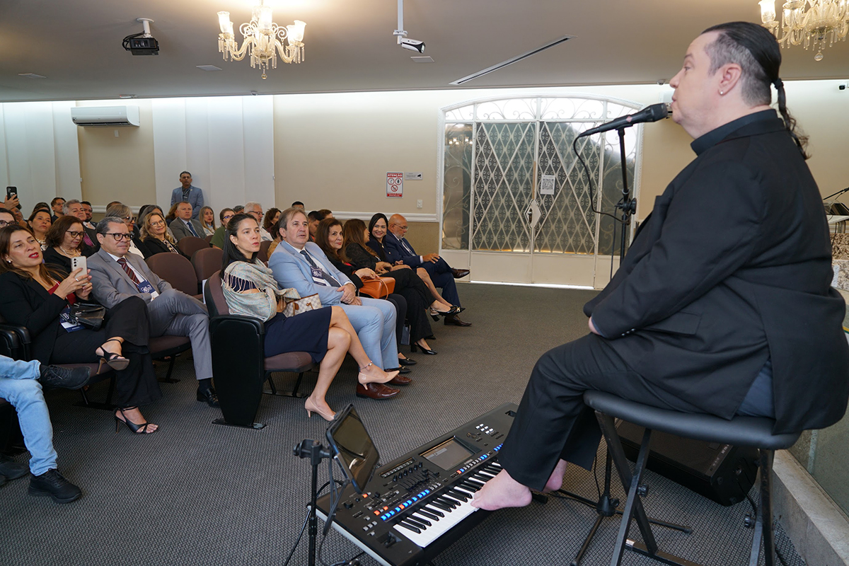 Uma cena de apresentação musical em um auditório. Em primeiro plano, no canto direito, um músico está sentado em um banco alto de costas para a câmera, vestindo um terno preto e com o cabelo preso. Ele está cantando em um microfone e seus pés descalços estão próximos a um teclado musical no chão. Em frente ao músico, há uma plateia de pessoas sentadas em cadeiras acolchoadas, observando a apresentação. O ambiente tem lustres no teto, e ao fundo, há uma grande porta de vidro com uma grade.