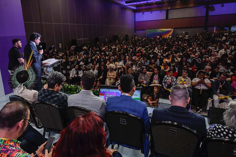 Uma foto panorâmica de um auditório lotado, onde um orador fala ao microfone em um púlpito, ao lado de outra pessoa com cocar. O público assiste atentamente, muitos usando acessórios coloridos.