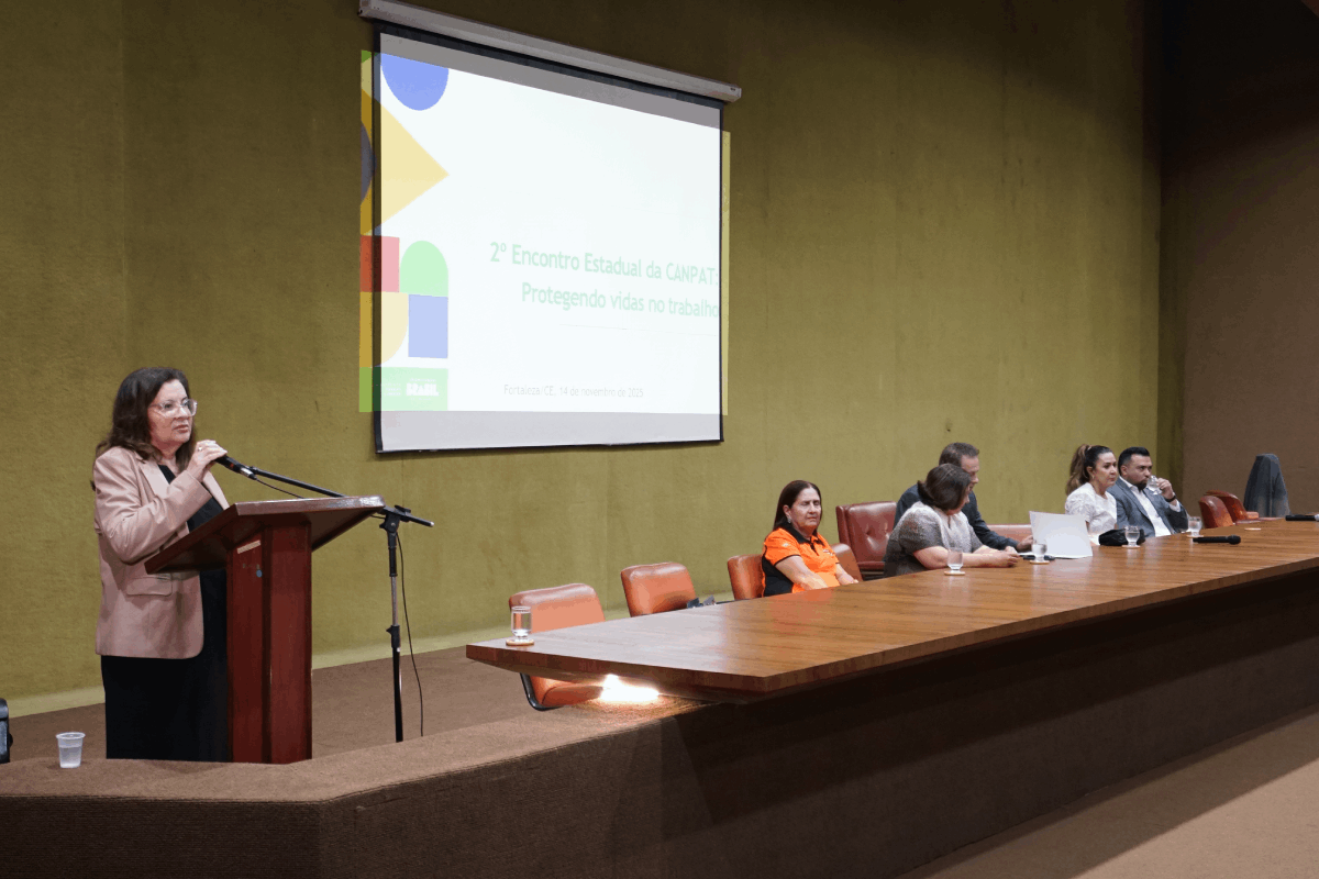 Uma mulher de meia-idade, com cabelos castanhos-escuros e óculos, está em pé em um púlpito de madeira escuro, segurando um microfone com a mão esquerda. Ela veste uma blusa social clara e uma saia preta. Sentados à mesa principal, à direita, estão outros participantes, e uma tela de projeção exibe o tema do evento.