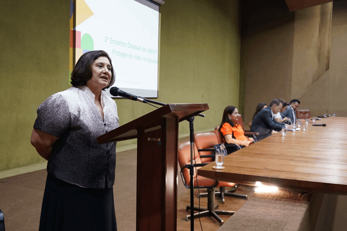 Uma mulher de meia-idade, com cabelos curtos e escuros, está em pé ao lado de um púlpito de madeira, falando ao microfone. Ela veste uma blusa com detalhes brilhantes na cor prata e preto e uma saia escura. Ao fundo, uma tela projeta o título do evento, e a mesa de debates com outros participantes se estende à direita.