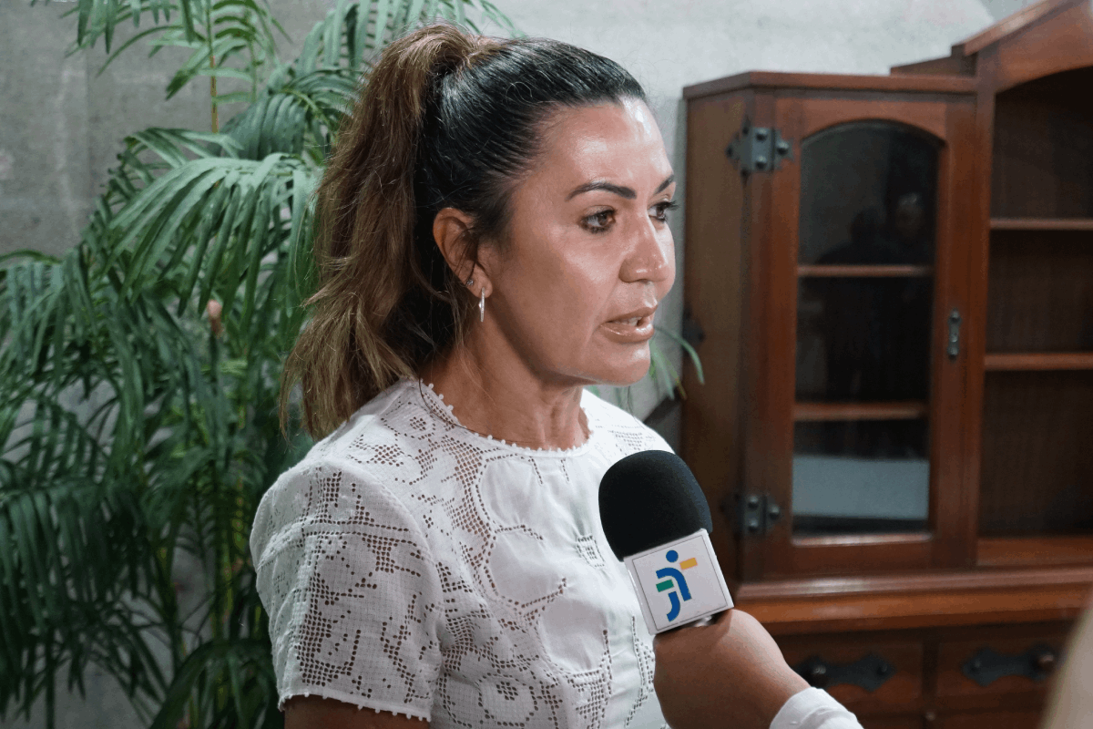 Uma mulher com pele bronzeada e cabelos escuros presos em um rabo de cavalo alto está em plano médio, falando em um microfone que tem um logotipo azul e verde. Ela veste uma blusa branca de renda com mangas curtas. Ao fundo, plantas grandes e um móvel de madeira escuro com prateleiras vazias completam a cena.