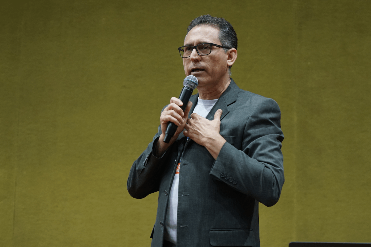 A imagem mostra um homem de meia-idade, com cabelos curtos grisalhos e óculos, em pé, segurando um microfone com a mão direita e com a mão esquerda sobre o peito. Ele veste um terno cinza escuro sobre uma camiseta branca. O fundo é uma parede lisa de tom verde-oliva.