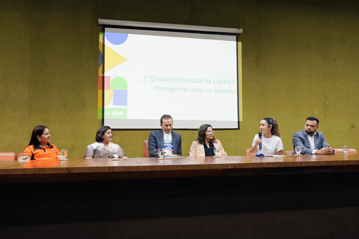 A imagem mostra seis pessoas sentadas em uma longa mesa de madeira em um auditório. Ao fundo, uma tela de projeção exibe a logo "2º Encontro Estadual da CANPAT: Protegendo vidas no trabalho". A maioria dos participantes veste roupas sociais, e uma mulher ao centro fala ao microfone, enquanto os demais a escutam atentamente.