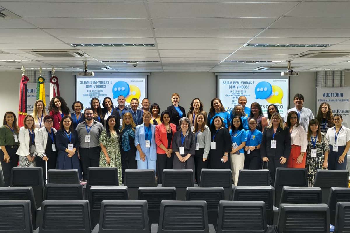Uma fotografia de um grupo de cerca de 30 pessoas, com uma mistura de homens e mulheres de diversas idades e etnias, posando para a câmera em um auditório. Eles estão em pé na frente de cadeiras vazias, alguns em grupos menores na frente, e atrás deles há duas telas de projeção com a frase "SEJAM BEM-VINDAS E BEM-VINDOS!" e o logotipo de um balão de fala.