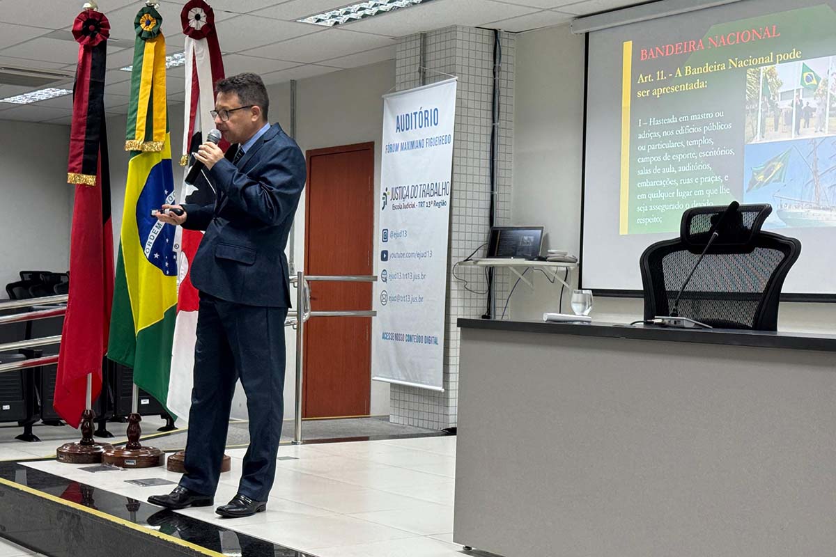 O servidor Antônio Carlos Santiago de Castro aparece em plano médio, no centro, vestindo um terno azul-marinho e ministrando o curso com um microfone na mão. À esquerda, são visíveis mastros com bandeiras (incluindo a Bandeira Nacional). À direita, uma tela de projeção exibe um slide sobre "Bandeira Nacional" e suas regras de uso.