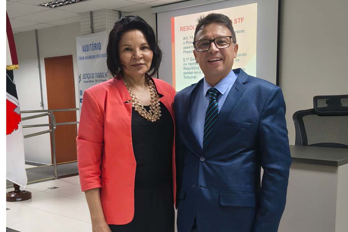 Fotografia em plano médio do instrutor Antônio Carlos Santiago de Castro, sorridente e de terno azul-marinho, posando ao lado de uma mulher. Ela veste uma jaqueta coral sobre uma roupa preta e um colar de pérolas volumoso. Eles estão em pé em frente a uma tela de projeção que exibe a palavra "RESOLUÇÃO" e referências a artigos sobre protocolo institucional.