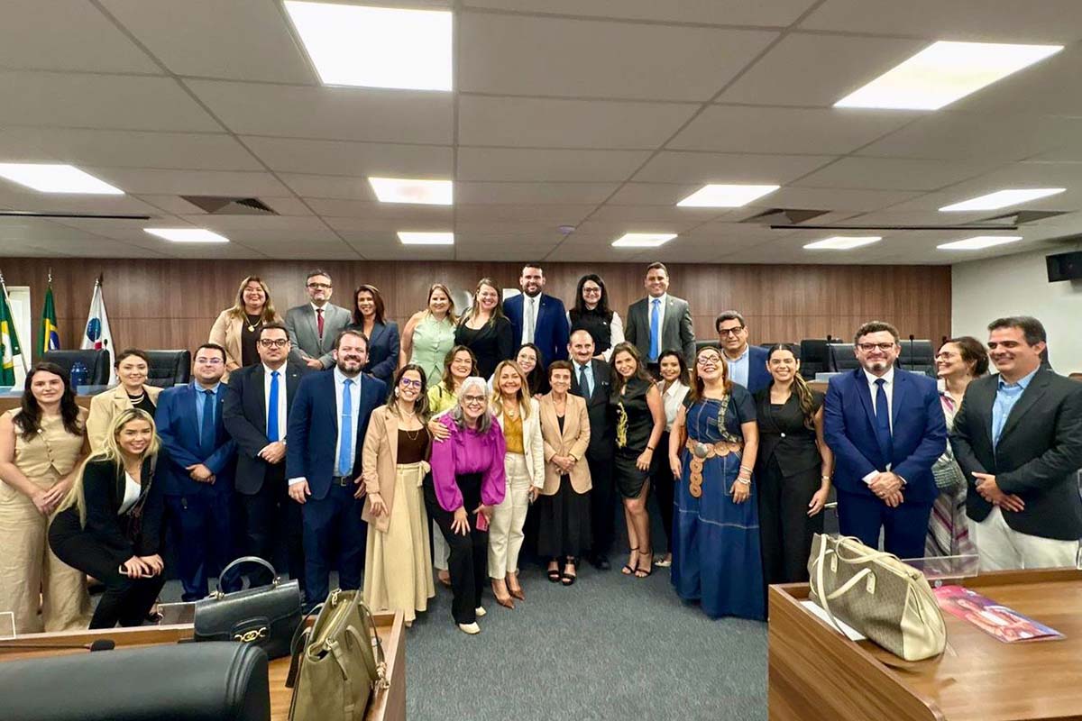 Um grupo de cerca de 25 advogados, homens e mulheres de diversas idades, posa de corpo inteiro em um auditório moderno. O grupo está organizado em três filas e sorri para a câmera. Ao fundo, uma parede de madeira escura e painéis de tecido exibe bandeiras do Brasil e do Ceará.