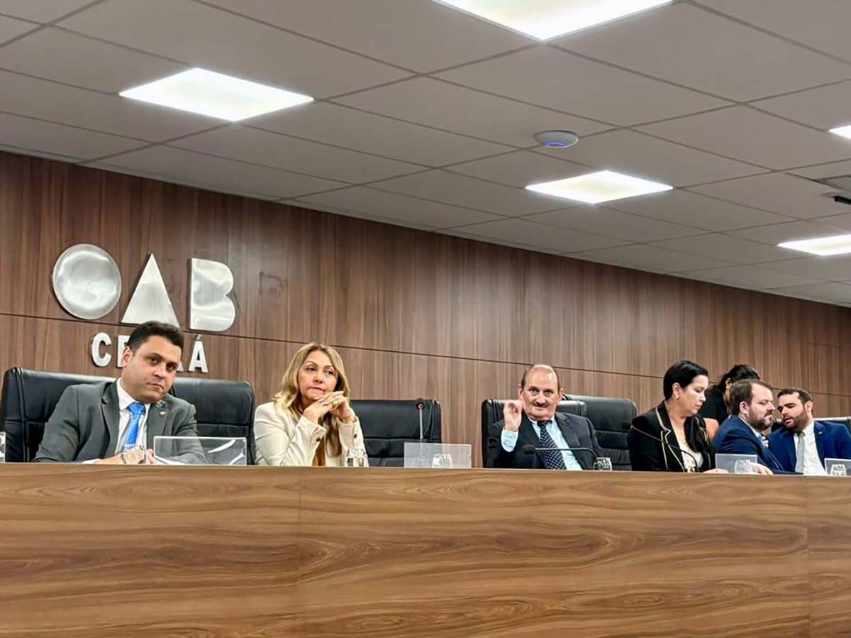 Quatro pessoas, duas mulheres e dois homens, sentados à mesa principal durante um evento, observam a audiência com atenção. Eles estão atrás de uma bancada de madeira escura e usam trajes sociais formais. Ao fundo, uma parede de madeira marrom exibe em relevo a logomarca da OAB Ceará.
