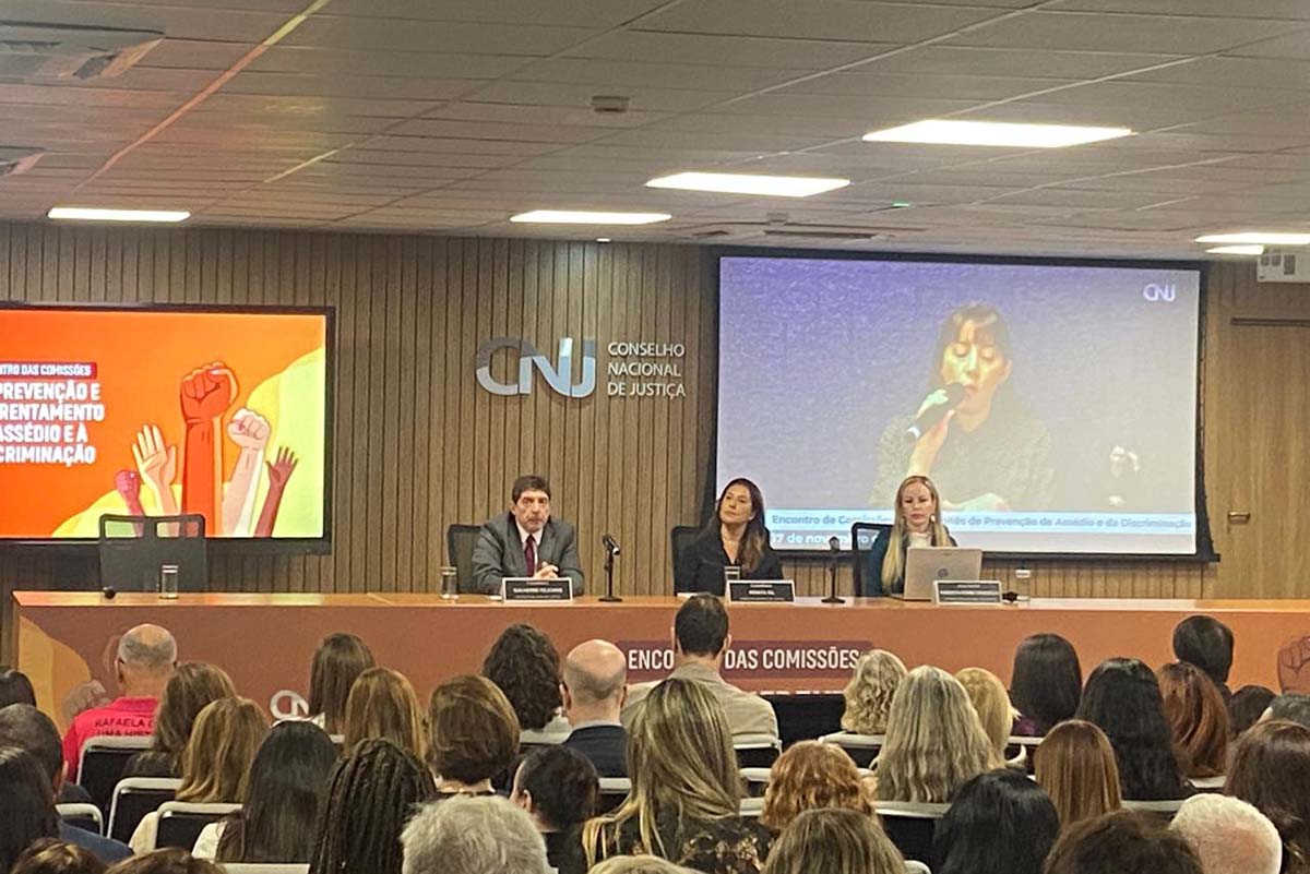 Uma foto de ambiente interno mostra uma mesa de painel composta por um homem e duas mulheres sentados à frente de um auditório lotado, de costas para a câmera. Ao fundo, um telão à direita exibe uma palestrante falando ao microfone, enquanto um segundo telão à esquerda mostra a identidade visual do evento: "Encontro das Comissões de Prevenção e Enfrentamento ao Assédio e à Discriminação". O painel principal traz o logotipo do Conselho Nacional de Justiça (CNJ).