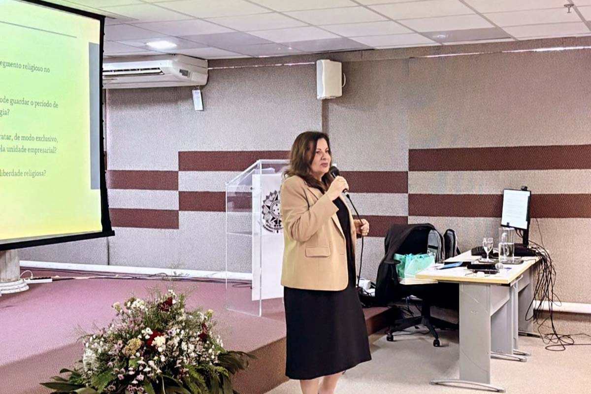 Uma mulher, com blazer bege e saia preta, fala segurando um microfone. Atrás dela, uma tela de projeção exibe o tema "liberdade religiosa" e há um púlpito de acrílico com o símbolo da República.