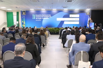 Uma foto registra um grande auditório lotado, visto de trás para frente. Dezenas de participantes, a maioria em trajes sociais, estão sentados em cadeiras em semicírculo, voltados para o palco. No palco, um telão azul de fundo mostra o título "IV Encontro Nacional dos Centros de Inteligência do Poder Judiciário", e um palestrante está discursando no púlpito à direita