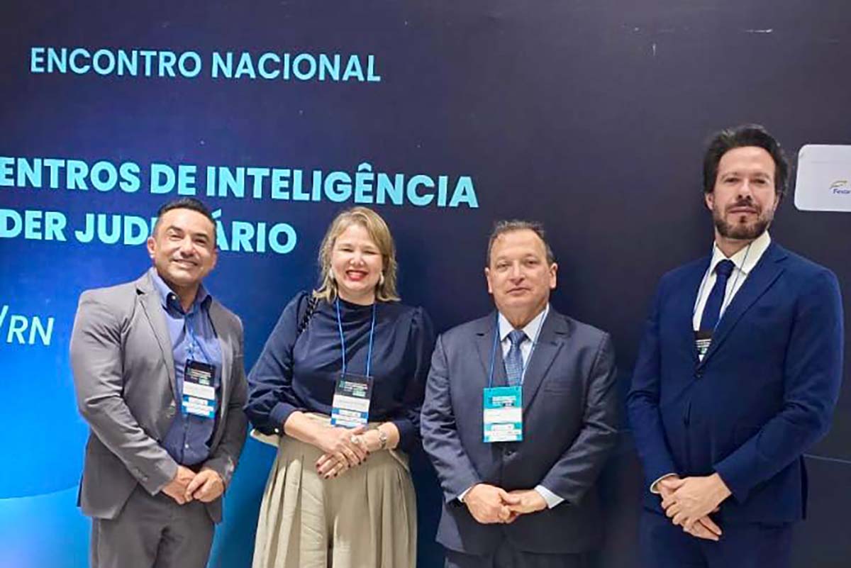 Uma fotografia de quatro pessoas, dois homens e duas mulheres, sorrindo para a câmera, em pé diante de um painel de evento. O painel, em azul escuro com texto branco, exibe "ENCONTRO NACIONAL" e "CENTROS DE INTELIGÊNCIA DO PODER JUDICIÁRIO". As pessoas usam crachás pendurados no pescoço.