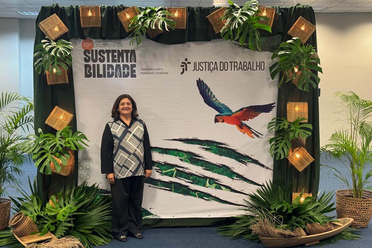 Uma mulher de meia-idade com blusa estampada em preto e branco e calças escuras está em pé no centro, sorrindo, em frente a um grande banner decorado. O banner branco, com detalhes verdes e uma arara em voo, anuncia o "10º Encontro de Sustentabilidade" e "Justiça do Trabalho". Ao redor, o cenário é complementado com folhagens tropicais, cestos de vime e luminárias suspensas, criando uma atmosfera natural.