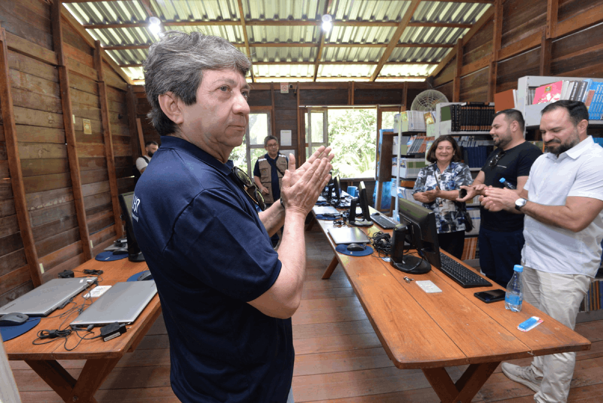 Uma foto de plano médio mostra um homem de meia-idade em primeiro plano, vestindo uma camisa polo azul-marinho, batendo palmas em um gesto de reconhecimento ou agradecimento. Ele está dentro de uma sala rústica, com paredes e teto de madeira, que funciona como um laboratório de informática. Ao fundo, outras quatro pessoas sorriem, e sobre as longas mesas de madeira, há monitores e notebooks doados