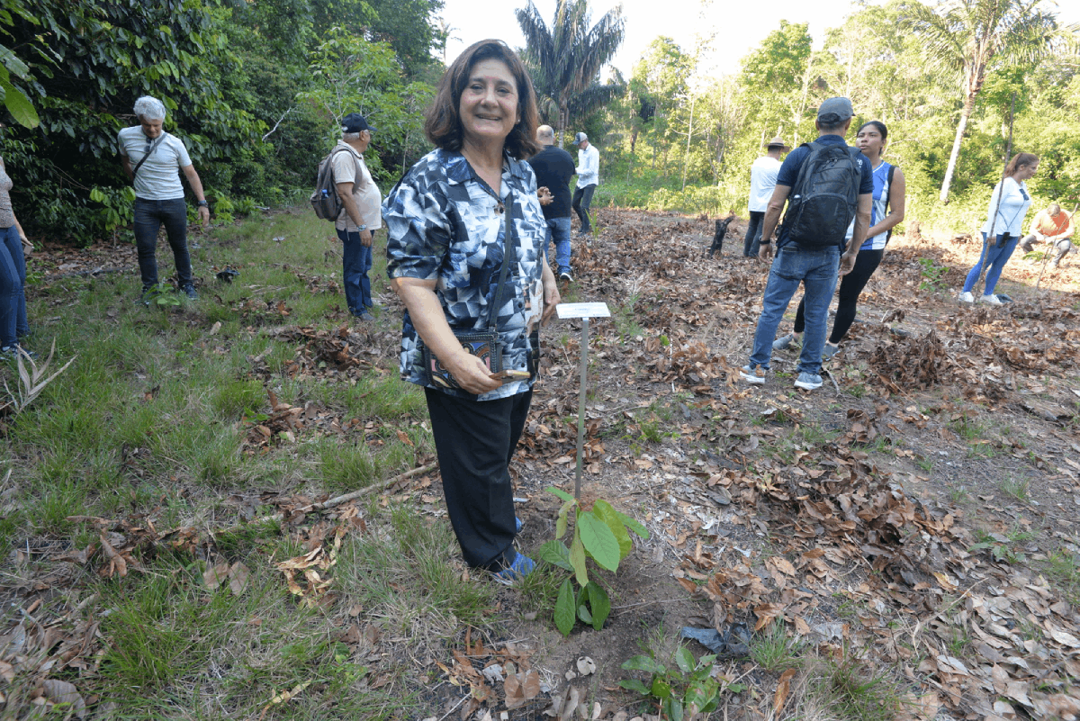 Uma mulher com blusa estampada em preto e branco e calça escura está em pé no centro da imagem, sorrindo para a câmera, ao lado de uma muda recém-plantada e uma pequena placa de identificação. Ela segura um smartphone em uma das mãos. Ao seu redor, o solo está coberto por folhas secas e há um grupo de pessoas ao fundo, algumas caminhando e outras observando, com uma vegetação densa compondo o cenário.