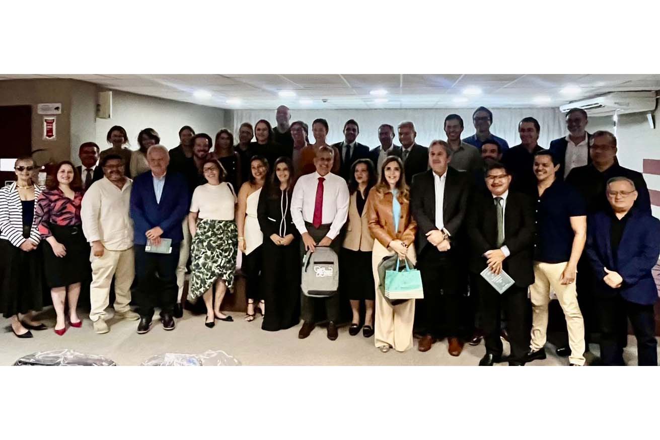 Um grande grupo de pessoas sorri para a câmera, em pé no auditório, após um evento. Há homens e mulheres, alguns com pastas ou sacolas, e um buquê de flores se destaca à frente. O ambiente possui paredes com faixas horizontais e um ar-condicionado visível.