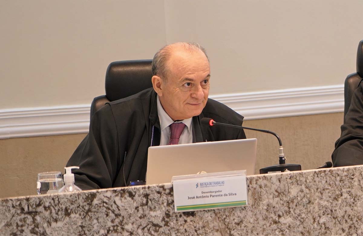  🎤 Audiodescrição e Legenda Audiodescrição (3 linhas) Um homem branco de meia-idade, careca e vestindo toga de magistrado, está sentado em uma mesa de conferência. Ele sorri levemente e olha para a direita, atrás de um notebook aberto. Sobre a mesa, há uma placa de identificação que o nomeia como Desembargador José Antônio Parente da Silva.