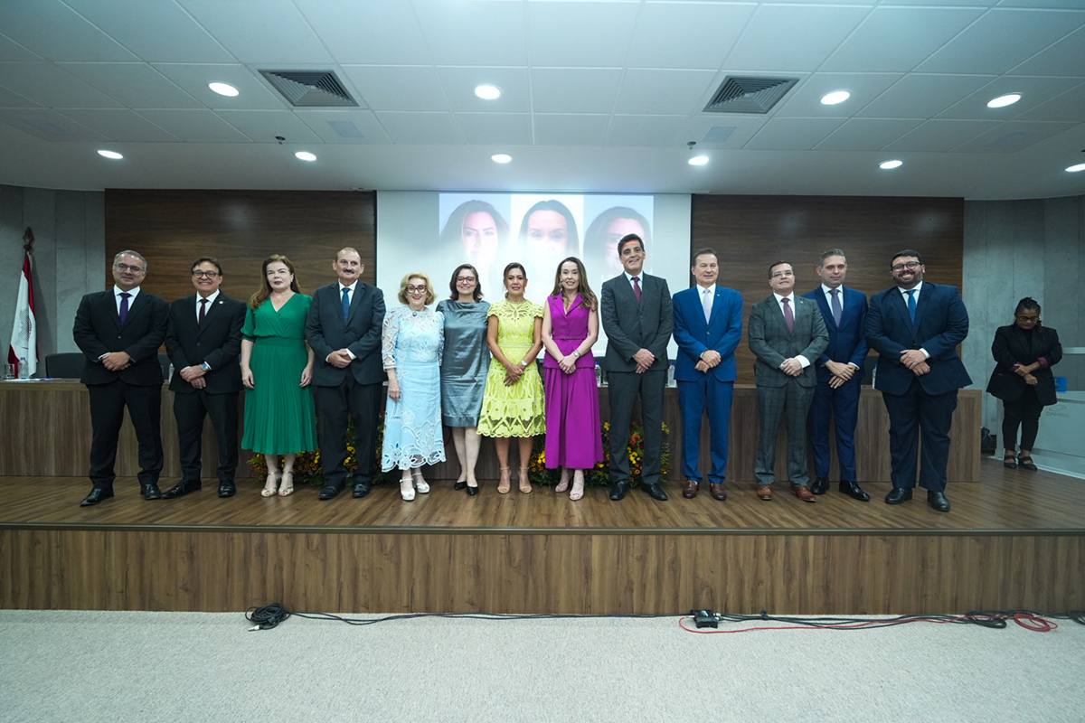Foto em plano médio mostra 13 pessoas de pé no palco, sobre um piso de madeira, durante uma solenidade. A maioria veste trajes sociais (ternos escuros) e há quatro mulheres em vestidos coloridos (verde, azul claro, amarelo e magenta).  Ao fundo, um telão exibe quatro rostos de mulheres, com destaque central para a nova procuradora-chefe, Ana Valéria Targino de Vasconcelos.  A mesa oficial, composta por autoridades, está posicionada na ordem da esquerda para a direita, celebrando a posse no Ministério Público do Trabalho no Ceará.