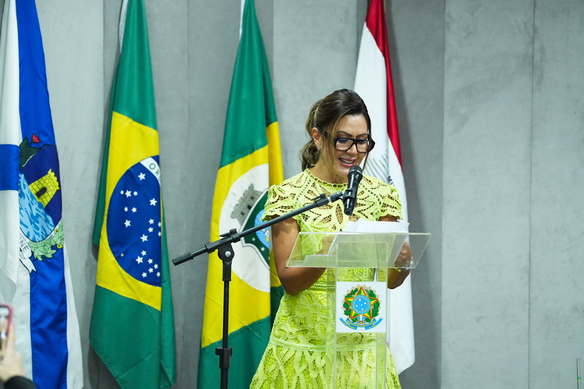 A imagem, em plano médio, focaliza a nova procuradora-chefe, Ana Valéria Targino de Vasconcelos, no momento do seu discurso. Ela veste um vestido verde limão de renda e usa óculos.  Ela sorri enquanto lê anotações em um púlpito transparente que exibe o brasão da República, com um microfone à sua frente.  Três bandeiras (duas do Brasil e uma do Ceará, com seus respectivos emblemas) e uma quarta bandeira (branca, vermelha e preta, provavelmente do MPT) servem de fundo, alinhadas à esquerda da empossada.