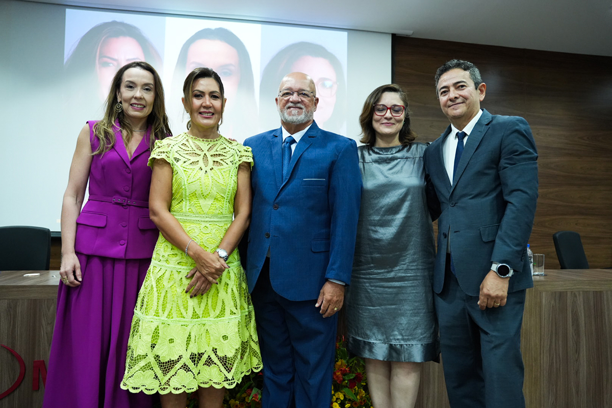 A imagem mostra cinco pessoas, três mulheres e dois homens, lado a lado em um ambiente interno.  Ao centro, Ana Valéria Targino de Vasconcelos, de vestido verde-limão, sorri entre as procuradoras Giselle Alves (à esquerda, de roxo) e Christiane Nogueira (à direita, de cinza), além de dois homens em ternos escuros.  Ao fundo, um telão exibe os rostos das três mulheres que compõem a nova gestão, com destaque para a nova procuradora-chefe.