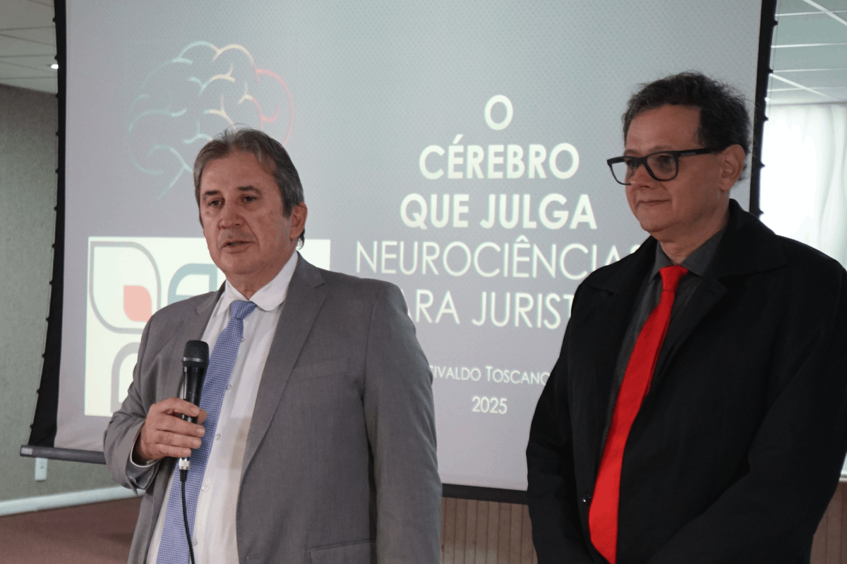 Foto em plano médio frontal de dois homens em pé, em frente a uma tela de projeção com o tema "O CÉREBRO QUE JULGA - NEUROCIÊNCIA PARA JURISTAS". À esquerda, o desembargador Paulo Régis Machado Botelho, de terno cinza claro e gravata azul, segura um microfone. À direita, o juiz Rosivaldo Toscano Júnior, de paletó preto e gravata vermelha, sorri discretamente, olhando para frente.