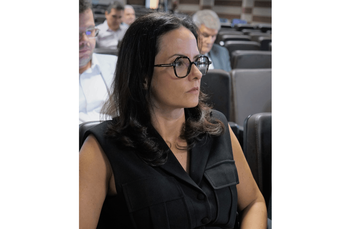 A foto em primeiro plano, na altura do busto, mostra a juíza do trabalho Rafaela Soares Fernandes atenta. Ela tem cabelos escuros e ondulados, usa óculos de armação geométrica preta e veste uma blusa preta de estilo colete. Ao fundo, embaçados, outros participantes sentados no auditório.