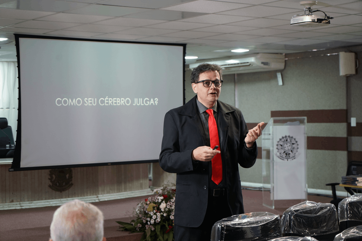 O juiz Rosivaldo Toscano Júnior, vestido de paletó preto e gravata vermelha, está em pé em um auditório, gesticulando enquanto profere sua palestra. Atrás dele, uma tela de projeção exibe em letras brancas a pergunta "COMO SEU CÉREBRO JULGA?". Em primeiro plano, parte da cabeça de um participante, vista de costas.