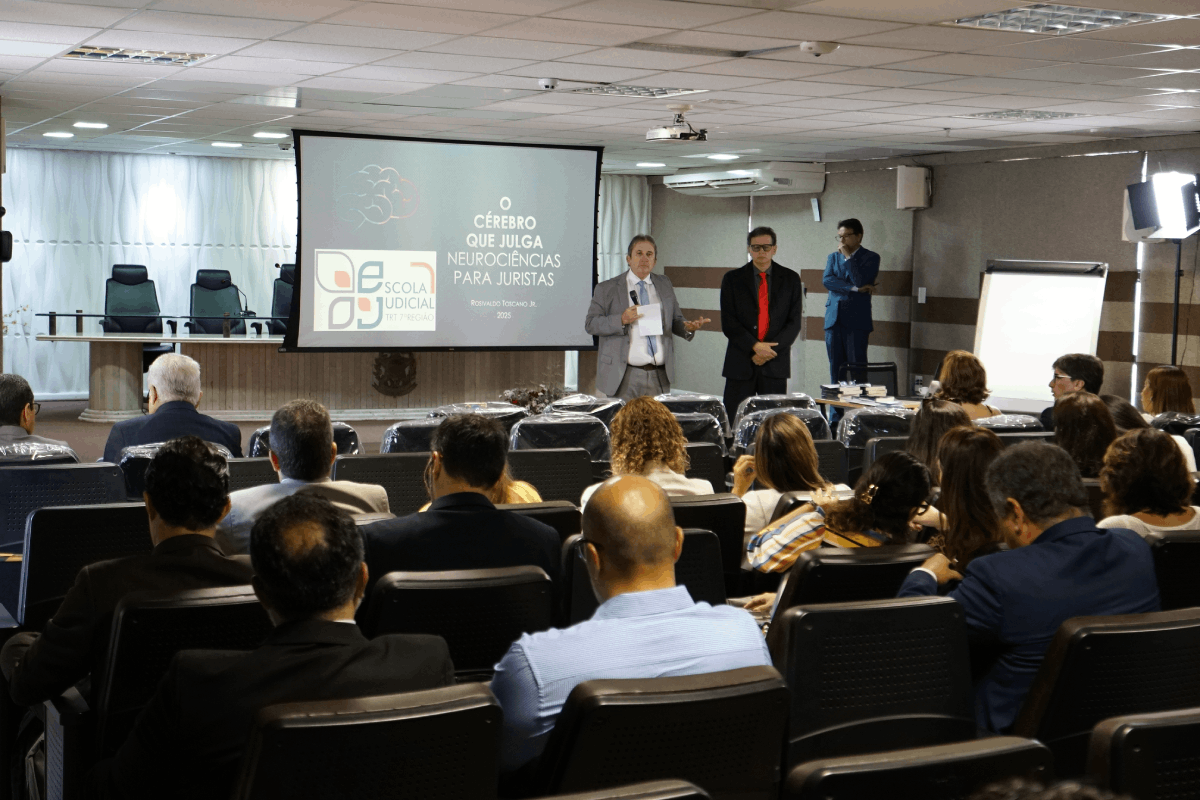 Uma foto registra o auditório da Escola Judicial do TRT-CE durante o Workshop "O cérebro que julga". O palestrante, à direita e de terno, fala ao microfone em frente a uma tela de projeção com o logotipo da EJUD e o tema Neurociências para Juristas. Vários magistrados assistem à palestra, vistos de costas, sentados nas primeiras filas.
