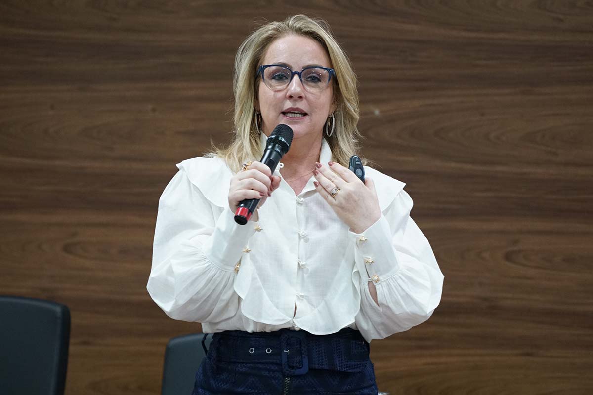 Uma mulher loira de óculos, vestindo blusa branca de mangas bufantes e saia azul escura, fala ao microfone. Ela gesticula com a mão esquerda, que exibe anéis, e tem a boca aberta em expressão de fala. Ao fundo, uma parede de madeira escura.