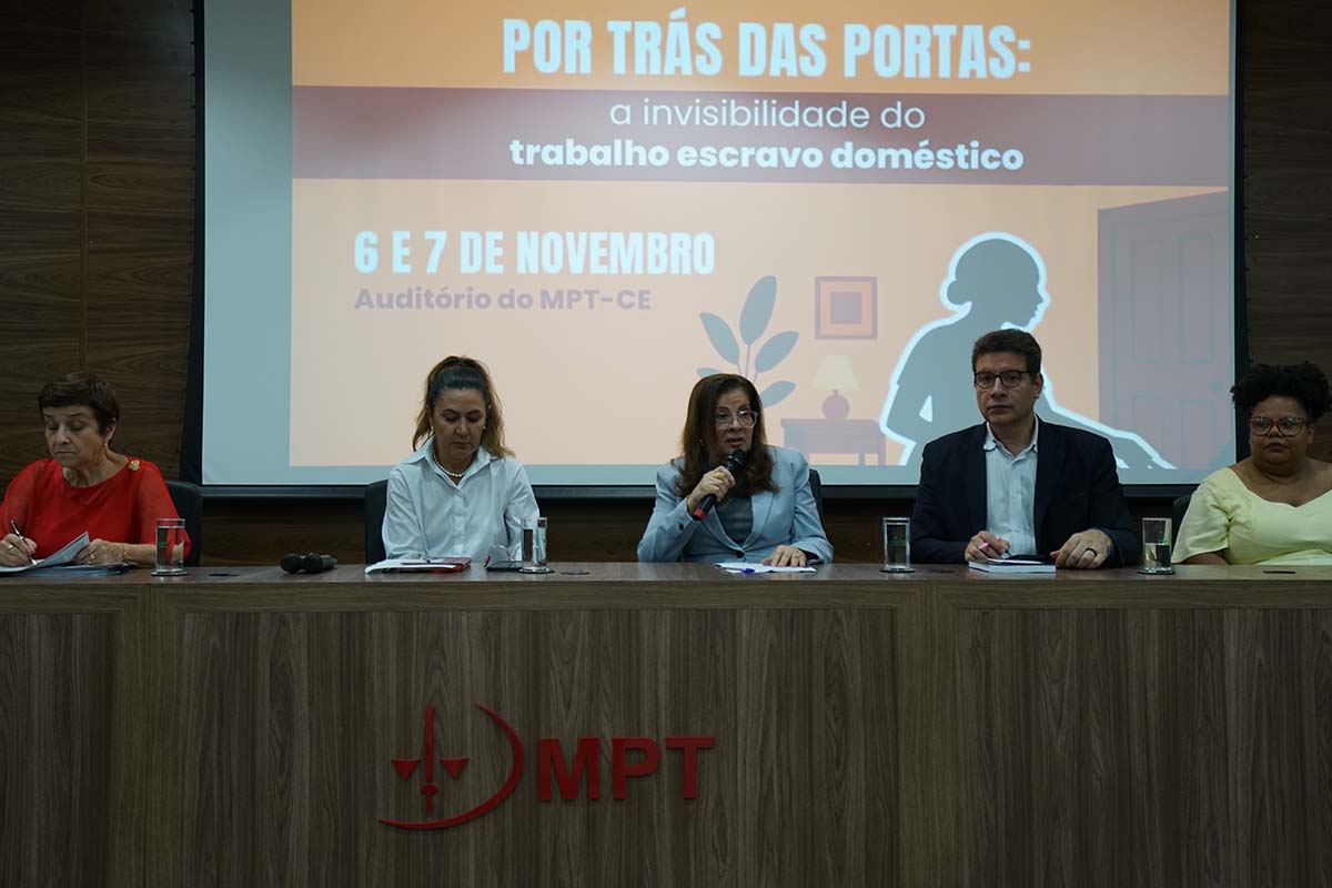 Uma foto frontal de cinco palestrantes sentados à mesa de debate, com a Presidente do TRT-CE, Desembargadora Fernanda Uchôa, no centro, falando ao microfone. O painel de fundo exibe o título do evento: "Por Trás das Portas: a invisibilidade do trabalho escravo doméstico" e as datas "6 e 7 de novembro" .
