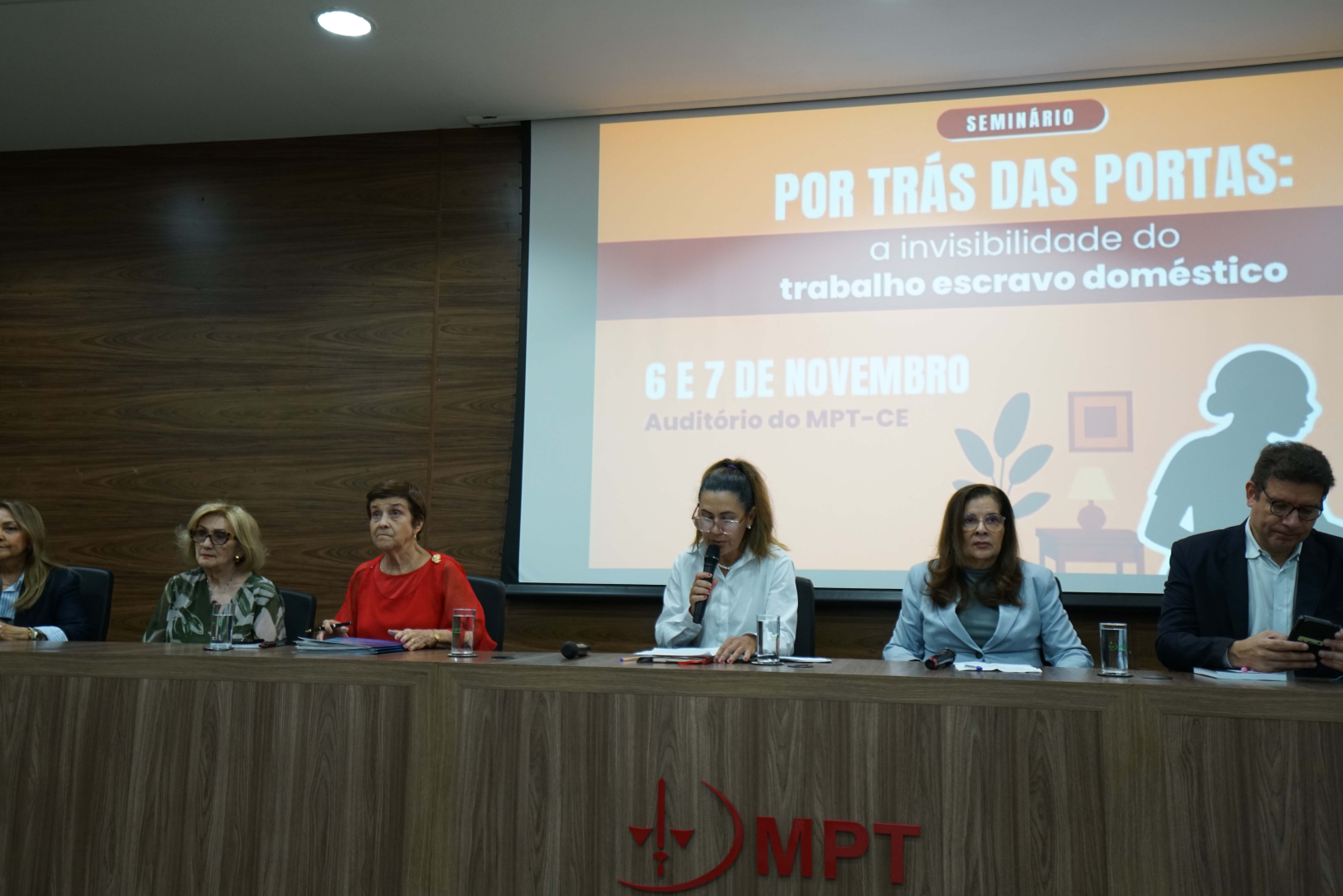 Uma vista frontal de seis autoridades sentadas à mesa de debate, com o telão de fundo exibindo o tema do seminário. A Procuradora-Chefe Ana Valéria Targino, de camisa branca, está ao centro falando ao microfone. A Ministra Delaíde, de blusa vermelha, e a Presidente do TRT-CE, Fernanda Uchôa, também compõem a mesa.