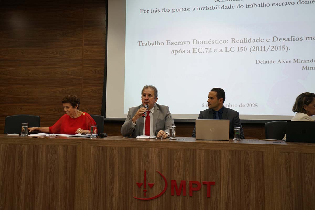 Um homem de terno cinza e gravata vermelha, o Desembargador Paulo Régis Machado Botelho, está no centro de uma mesa escura, falando ao microfone . Ele está ladeado pela Ministra Delaíde Alves Miranda, de blusa vermelha, à esquerda. O telão ao fundo exibe o título da conferência.