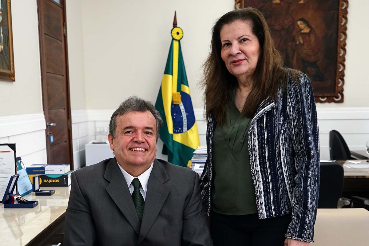 Um homem de cabelos grisalhos e terno cinza sorri, sentado em primeiro plano. Atrás dele, à direita, uma mulher de cabelos escuros e jaqueta listrada sorri, em pé. Ao fundo, uma bandeira do Brasil e um quadro em moldura dourada.