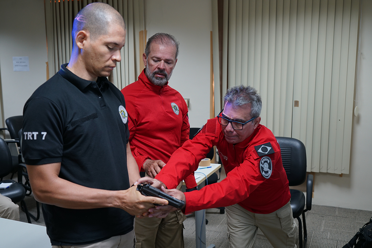 Um ambiente interno com cortinas bege e cadeiras pretas. Três homens participam de um treinamento: um jovem calvo, com uma camisa polo preta e o nome "TRT 7" na manga esquerda, segura uma pistola com as duas mãos. À sua frente e à direita, um instrutor mais velho, de camisa vermelha, óculos e calças bege, está agachado e o auxilia ativamente, colocando as mãos sobre a arma para demonstrar uma técnica. Um terceiro homem, também de camisa vermelha, barba grisalha e calças bege, observa a instrução entre os outros dois.