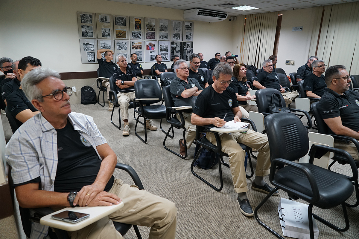 Uma sala de aula, com carpete e paredes brancas decoradas com fotos antigas em preto e branco. Cerca de vinte pessoas, predominantemente homens de meia-idade ou mais velhos, estão sentadas em cadeiras pretas e dispostas em fileiras, assistindo a uma palestra ou aula. A maioria usa camisas pretas do treinamento e calças beges, e alguns tiram notas em cadernos. Em primeiro plano, à esquerda, um homem de camisa quadriculada clara sobre a camisa preta usa óculos e observa a frente da sala.
