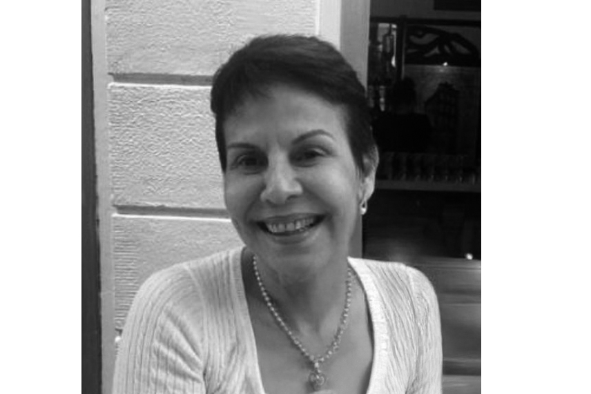 Uma foto em preto e branco de uma mulher de meia-idade, com cabelo curto e escuro, sorrindo gentilmente. Ela usa um cardigã claro, uma corrente de pérolas e um brinco pequeno. O fundo é uma parede de pedra ou azulejos claros.