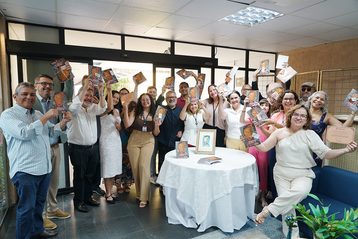 Um grupo diversificado de cerca de 20 pessoas celebra em um ambiente interno bem iluminado, segurando livros com a mesma capa abstrata.  A maioria está de pé, sorrindo e levantando os livros no ar. No centro, há uma mesa redonda coberta com uma toalha branca, com uma foto emoldurada e cópias do livro no centro.  Os participantes usam roupas casuais e sociais, em cores claras e neutras.  Uma mulher na frente, à direita, se destaca, vestida com um macacão creme de ombro único, sorrindo e gesticulando amplamente com os braços abertos.  A celebração parece ser um lançamento ou evento literário, com uma porta de vidro ao fundo.