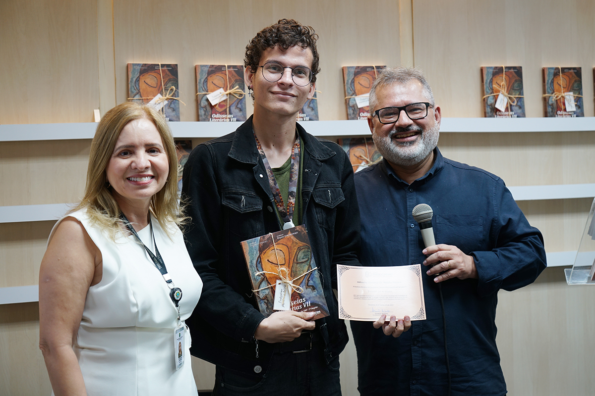 Três pessoas sorridentes posam em frente a uma prateleira com cópias de um livro.  À esquerda, uma mulher de cabelos loiros, vestindo um colete branco sem mangas. No centro, um jovem alto, de óculos e jaqueta jeans preta, segura o livro, cuja capa tem uma pintura abstrata em tons de laranja e azul.  À direita, um homem de barba grisalha e camisa azul escura segura um microfone e um certificado.  Atrás deles, várias cópias do livro, empilhadas horizontalmente, preenchem a prateleira.