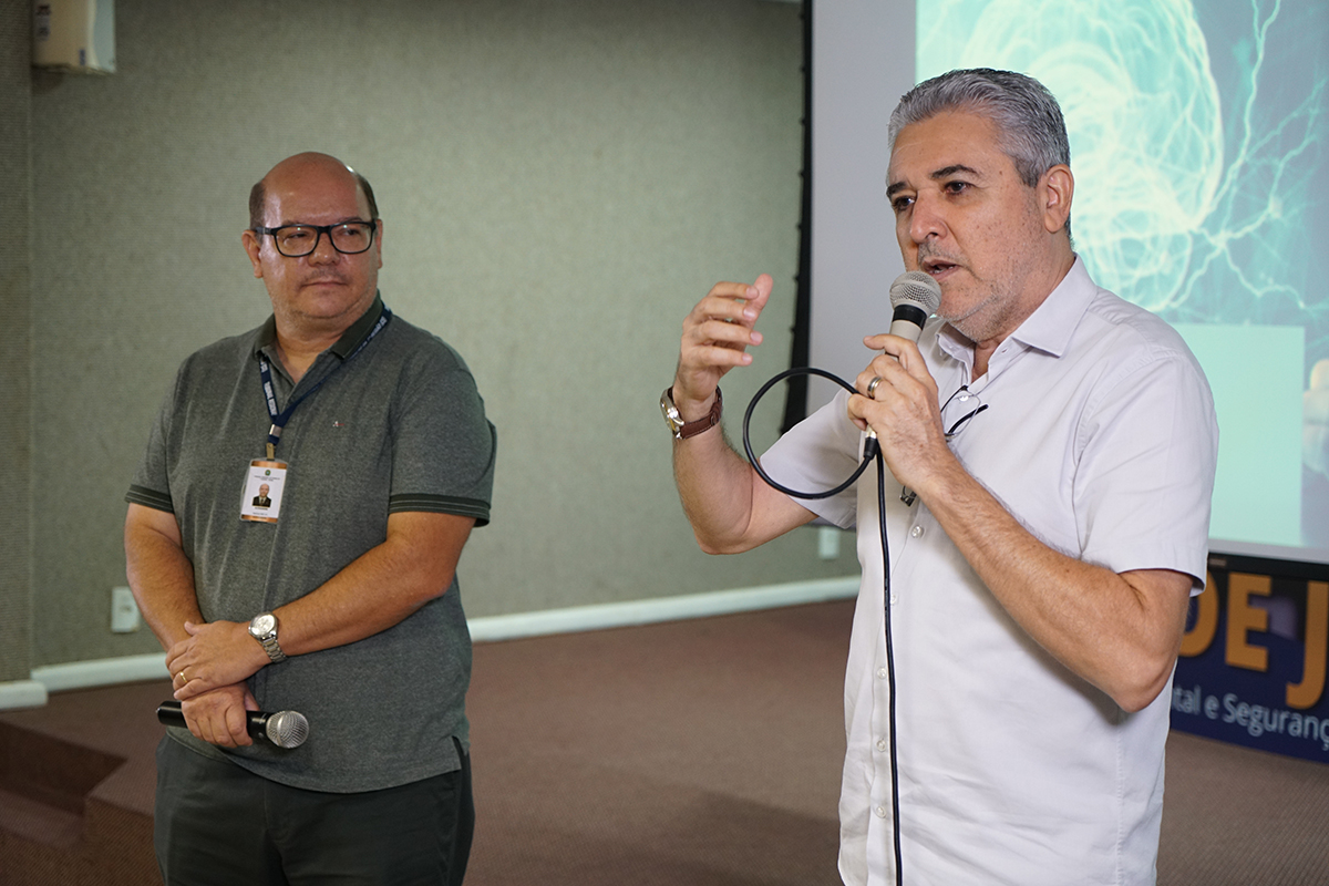 Dois homens de meia-idade estão em um palco ou área de apresentação, com uma tela de projeção atrás. À direita, um homem de cabelo grisalho e camisa social branca está falando em um microfone com fio, gesticulando com a mão direita. À esquerda, um homem calvo de óculos e camisa polo cinza escura segura outro microfone na frente do corpo com as duas mãos, olhando para a frente.