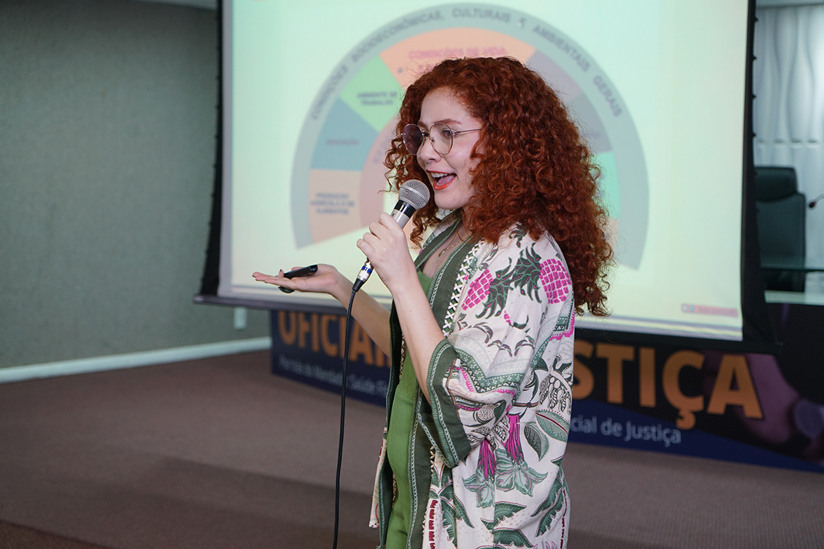 Uma mulher de cabelo ruivo cacheado e óculos redondos está no palco, falando e gesticulando com a mão aberta enquanto segura um microfone com fio na outra. Ela veste uma blusa verde sob um quimono branco com estampa de folhas verdes e flores vermelhas. Ao fundo, uma tela de projeção exibe um gráfico circular colorido na parte superior, e na inferior, palavras como "OFICIAIS" e "JUSTIÇA" em letras grandes.