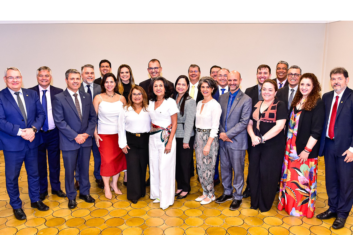 Uma foto de grupo com 19 pessoas (homens e mulheres) em trajes sociais, posando lado a lado contra uma parede clara e lisa. A iluminação é uniforme e o chão é coberto por um piso de mosaico em tons de amarelo e bege.  O grupo está bem alinhado, com a maioria dos homens vestindo ternos escuros (azul-marinho, cinza e preto). As mulheres estão vestidas com roupas de cores claras (branco, bege) e algumas estampas vibrantes, como a mulher na extrema direita com um vestido floral.  A atmosfera é formal e amigável, e todos no grupo estão olhando diretamente para a câmera, com sorrisos discretos ou expressões neutras.