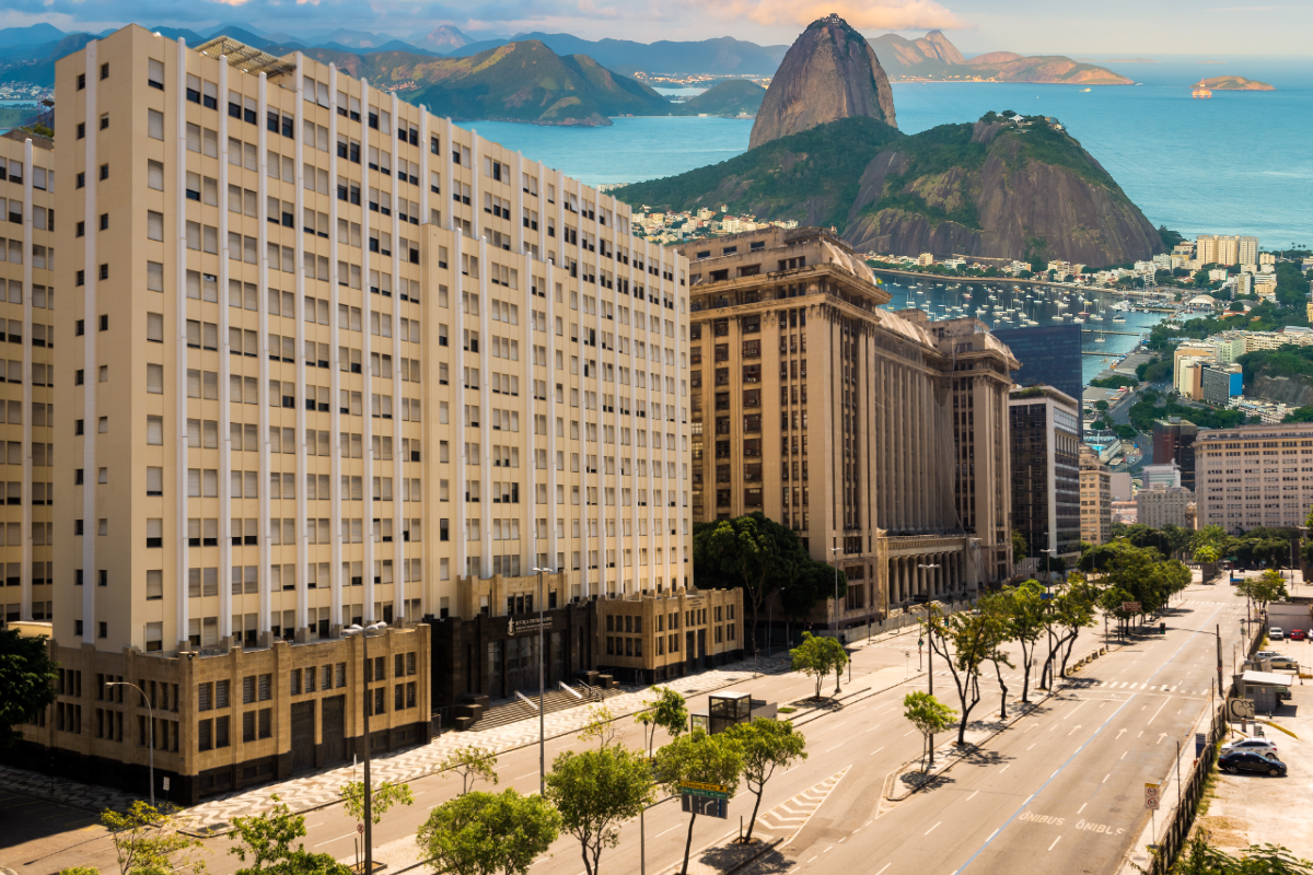  Fotografia panorâmica do centro do Rio de Janeiro sob luz solar. No primeiro plano, à esquerda, destaca-se o imponente edifício do TRT-RJ, em tons de bege, ladeando uma larga avenida arborizada e quase vazia. Ao fundo, a Baía de Guanabara exibe águas azuis repletas de barcos, emoldurada pelo icônico Pão de Açúcar e montanhas sob um céu levemente nublado.