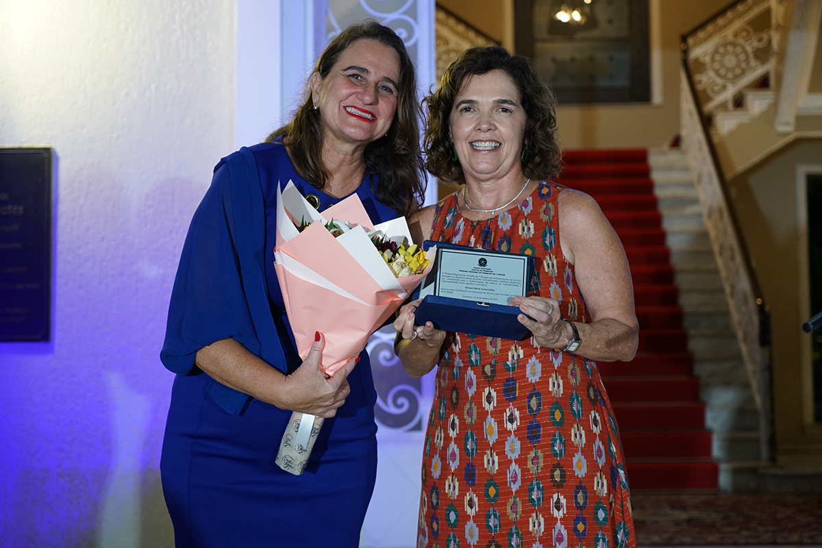 Foto da Assessora de Governança Deven Miller e a servidora homenageada Silvana Dias. Deven está entregando um buquê de rosas à Silvana que está segurando uma placa de homenagem.