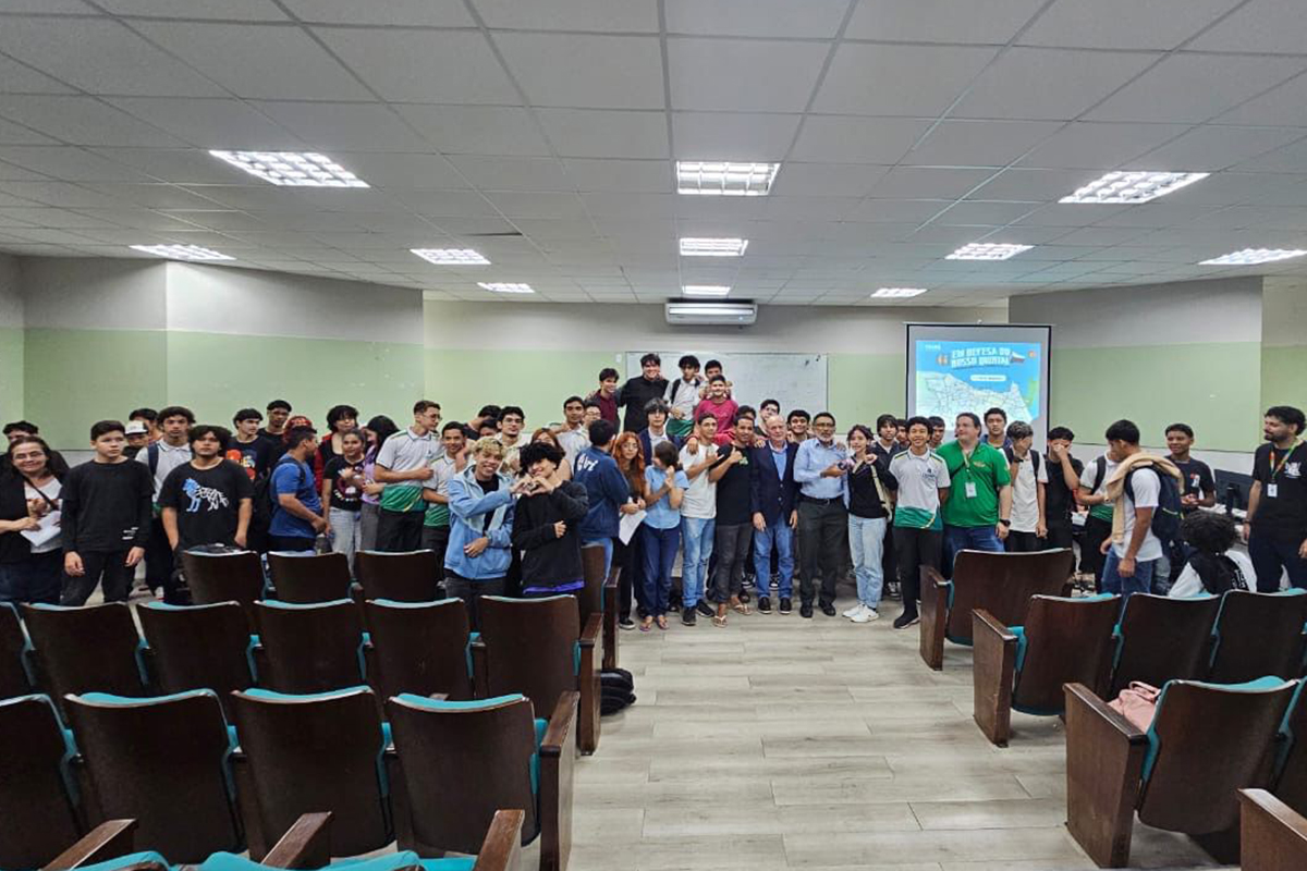 Uma foto de plano geral mostra um auditório iluminado com cadeiras vazias em primeiro plano. Um grande grupo de jovens, a maioria rapazes, está reunido no centro da sala, olhando para a câmera e parecendo celebrar um evento. Ao fundo, uma tela de projeção exibe um gráfico e texto.