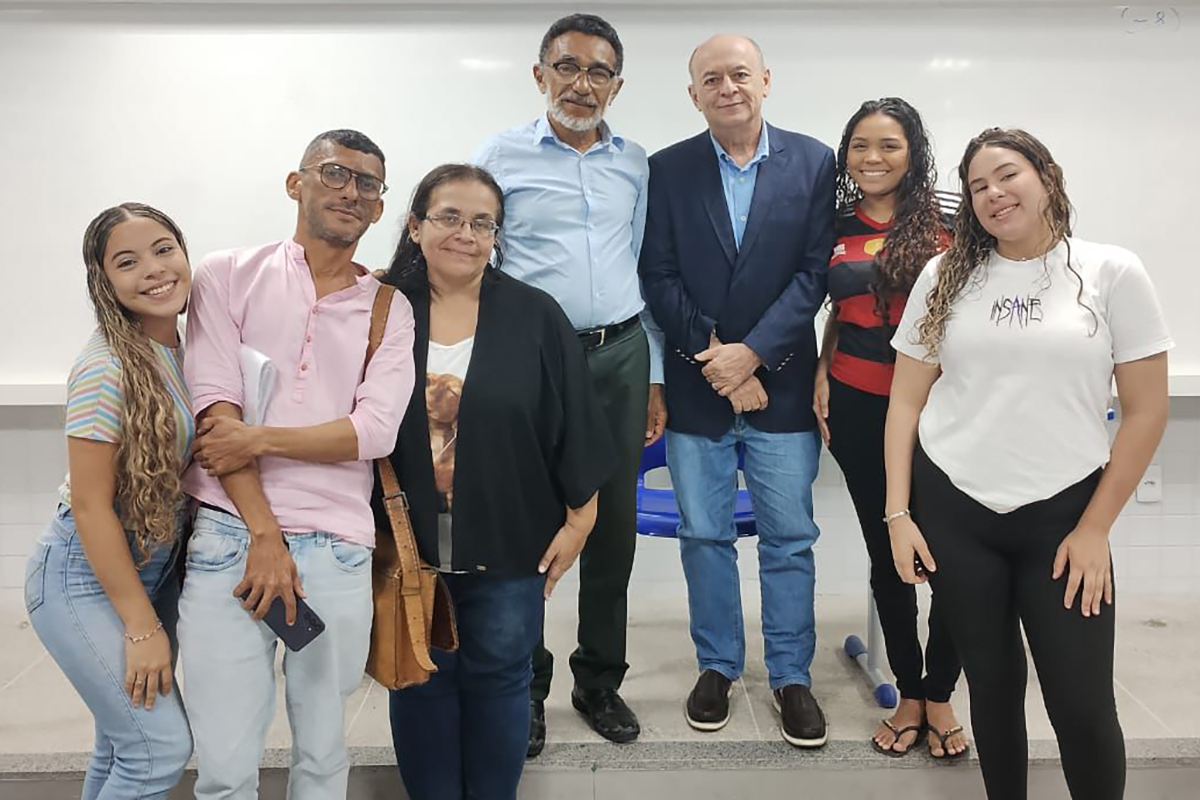 Uma foto de grupo mostra sete pessoas, quatro mulheres e três homens, em pé em um ambiente interno. Eles estão em frente a um grande quadro branco, sorrindo levemente para a câmera. O grupo parece amigável, com alguns braços dados, em uma sala de aula ou auditório.