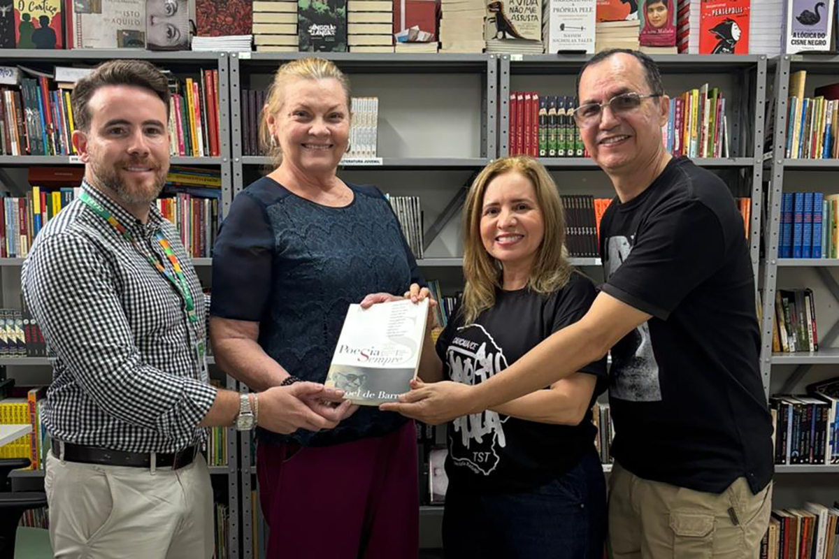  Quatro pessoas em pé, em frente a uma estante cheia de livros, participam de uma entrega. A bibliotecária Rejane Albuquerque, do TRT-CE (no centro-direita, com uma camisa preta com logo TST) entrega livros para o Chefe de Gabinete da SEAS, Artênio Igor de Oliveira Negreiros (primeiro à esquerda), e a bibliotecária da SEAS, Ana Dourado (segunda à esquerda). Sobre a mesa, em primeiro plano, estão pilhas de livros de literatura, história e cultura.