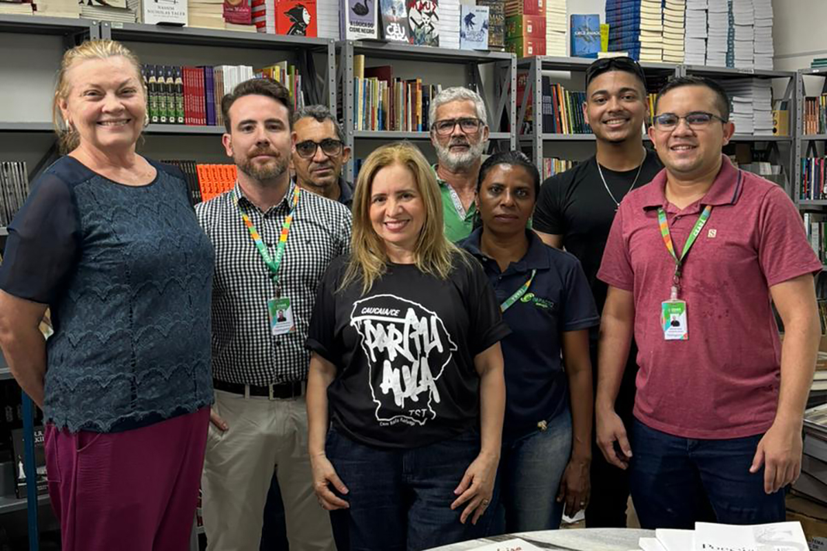 Quatro pessoas em pé, em frente a uma estante cheia de livros, participam de uma entrega. A bibliotecária Rejane Albuquerque, do TRT-CE (no centro-direita, com uma camisa preta com logo TST), entrega um livro a Artênio Igor de Oliveira Negreiros (primeiro à esquerda), Chefe de Gabinete da SEAS, e à bibliotecária da SEAS, Ana Dourado (segunda à esquerda), com um homem sorrindo à direita. Sobre a mesa, em primeiro plano, estão pilhas de livros de literatura, história e cultura.