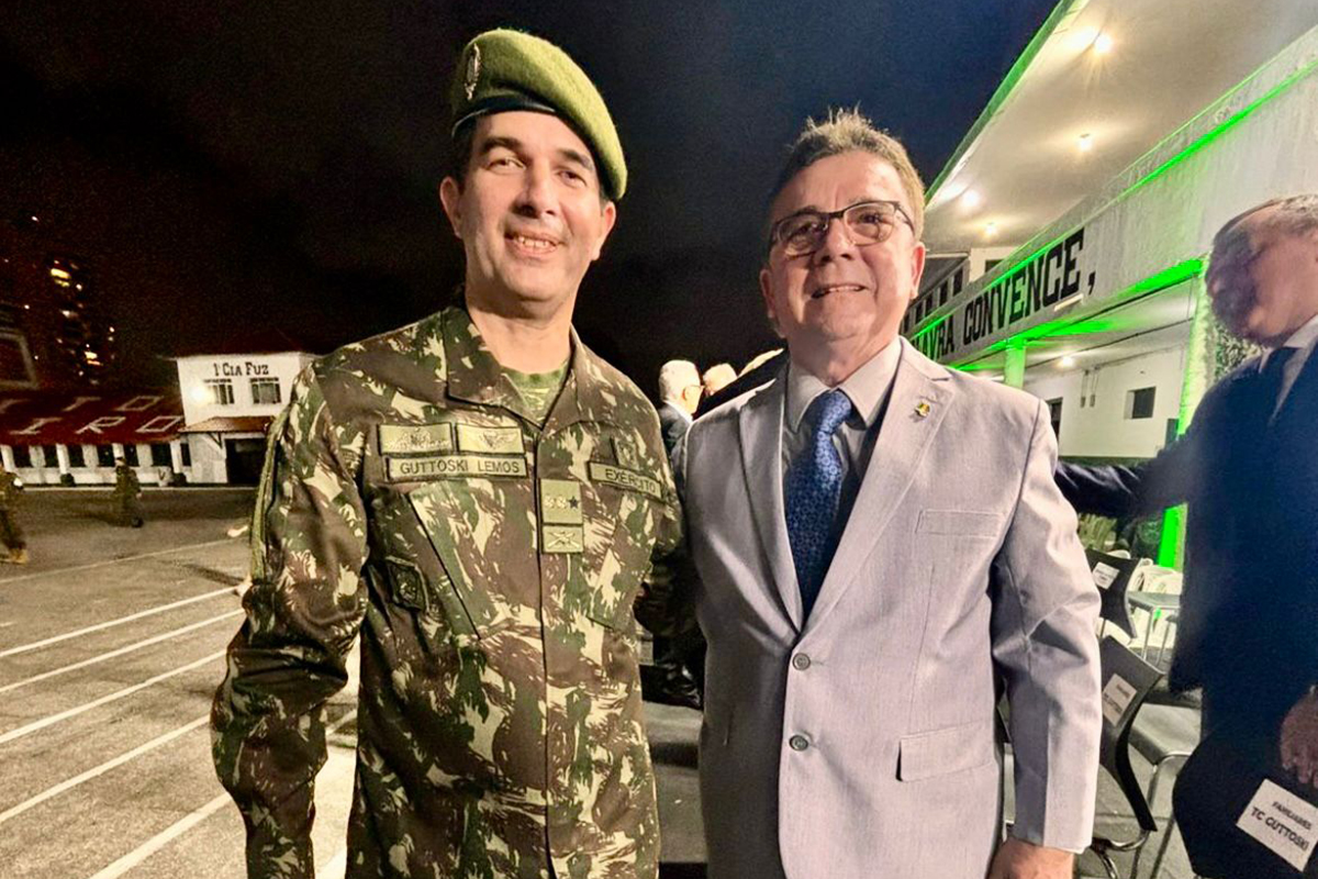 O Comandante Guttoski Lemos, em uniforme camuflado do Exército Brasileiro e boina verde, está à esquerda, sorrindo, ao lado de um civil.  O civil, à direita, veste um terno cinza claro, gravata azul e óculos, e também sorri para a câmera.  Ambos estão em uma área externa à noite, com iluminação verde sobre uma estrutura atrás deles, onde se lê parte de "CONVENÇÃO" (ou similar).