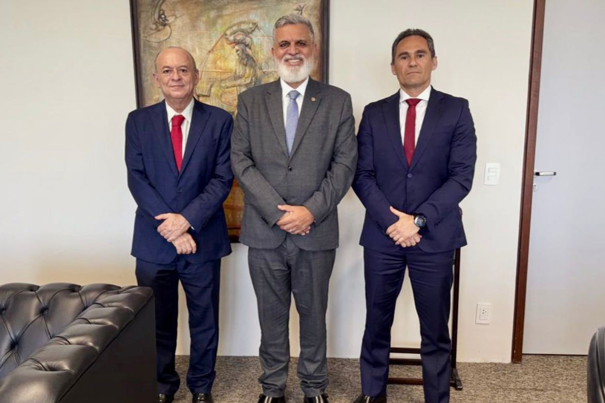 Três homens de pele clara e cabelos curtos, vestindo ternos formais e gravatas, posam de pé, lado a lado, em um ambiente interno. O homem ao centro tem barba branca e usa terno cinza, enquanto os outros dois vestem azul-marinho e gravatas vermelhas. Eles estão com as mãos cruzadas à frente do corpo, sorrindo discretamente para a câmera. Ao fundo, vê-se uma parede branca com um quadro abstrato e parte de um sofá de couro preto à esquerda.